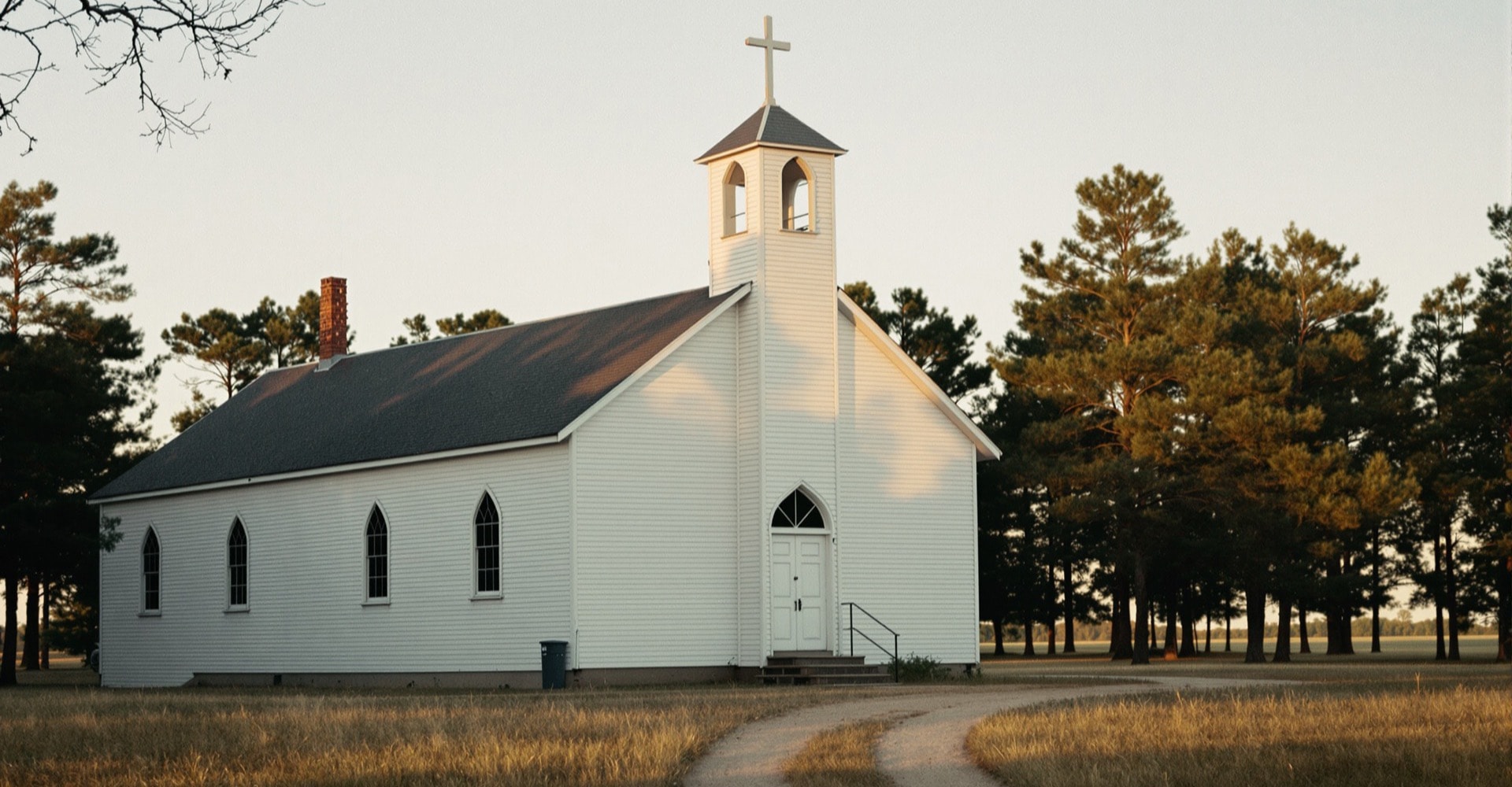 A white nineteenth-century timber church with a plain steeple standing among East Texas pines in late afternoon light